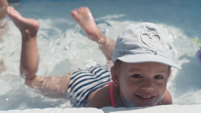 little happy 3 year old girl in blue cap and swimsuit Caucasian European Slavic appearance bathe and frolic in pool in summer, splashing water, lifestyle design. Activity nature leisure. Slow motion