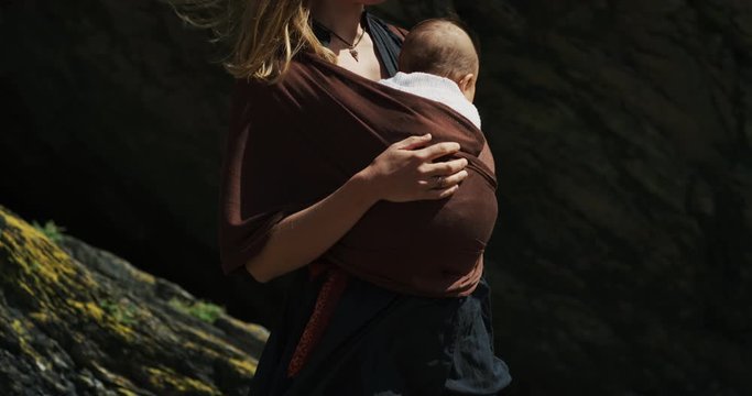Young mother withe baby in sling walking near cliff on beach