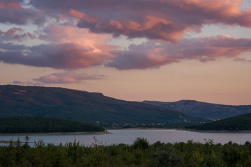 Naklejka na ściany i meble Pink clouds over the lake and forest. Bright sunset light. Purple and crimson shades. Rural landscape at sunset. A rustic motif with a lake and mountains.