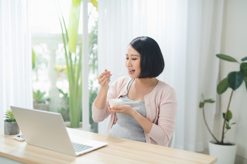 smiling redhead pregnant woman eating yoghurt while sitting at table with laptop at home