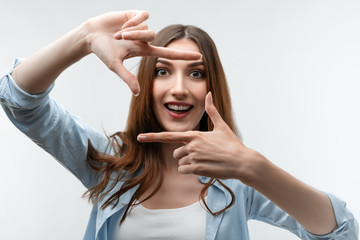 Positive girl with long chestnut hair gestures finger frame actively at camera.Human emotions, facial expression concept