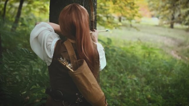 A woman archer hides near a tree, aims an arrow from a bow. Medieval girl hunter in a dense green forest. Сostume of a vintage warrior with a weapon in his hands. Fantasy shooting from back, rear view