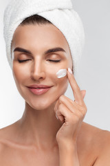 Close up portrait of half naked young woman applying moisturizing face anti-aging cream on flawless skin, closed eyes, isolated on grey background. Natural makeup, touching face. Beauty routine