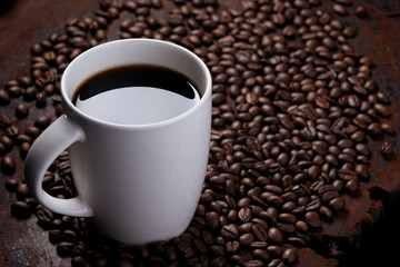 black coffee in white mug with surrounded by roasted coffee beans on dark red wood table