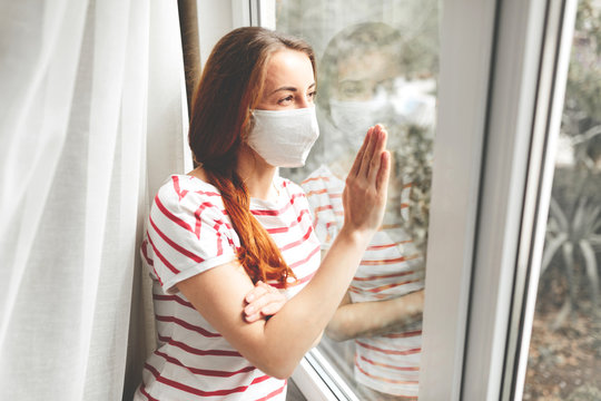 Outbreak concept. Portrait of a sad pensive young woman in medical mask in quarantine near the window.