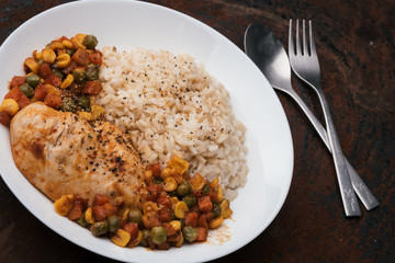 a dish of homemade roasted chicken breast and dice carrots, beans, corns with brown rice on dark red wooden table and cutlery