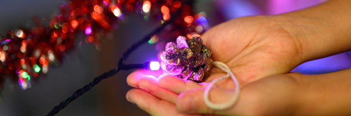 Christmas homemade DIY craft toy for the Christmas tree a cone with glitters in kids hands and New Year lights glowing garlands. banner