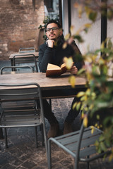Pensive caucasian man in glasses sitting at the table outdoors, holding open book in hands and looking at camera. Education concept