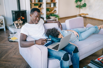 Black man with credit card and laptop sitting on sofa with sleeping woman