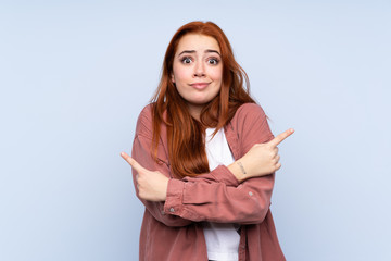 Redhead teenager girl over isolated blue background pointing to the laterals having doubts