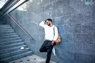 Portrait of stylish handsome man with a beard dressed white t-shirt, black sunglasses,  jeans with bagpack posing against the urban wall in the city on the street