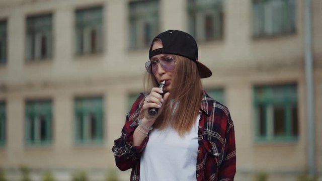 Pretty young hipster woman in black cap vape ecig. Girl vape popular ecig gadget, vaping device.Happy brunette vaper girl with e-cig. Portrait of smoker female model with electronic cigarette