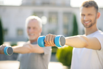 Fototapeta na wymiar Young bearded male and grey-haired man practicing with blue dumbbells outside, smiling