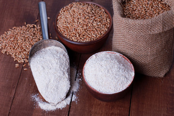 Bowl and scoop with wholegrain flour powder, sack of wheat grains on brown wooden table. High angle, closeup. Harvest, food, ingredient, baking concept