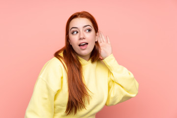 Redhead teenager girl over isolated pink background listening to something by putting hand on the ear