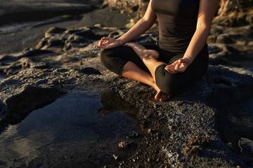Woman practicing yoga and meditation in lotus position on black stones near the sea.