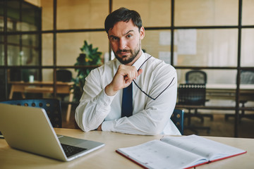 Portrait of Caucasian suspicious employee with optical glasses in hand looking at camera during work time in office interior, serious bearded businessman sitting at desk with computer technology