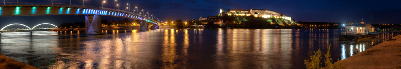 Petrovaradin fortress in Novi Sad, Serbia and Rainbow bridge illuminated with colorful street lights and reflection in the Danube river water, panorama shot