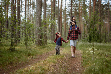 Obraz premium A father is walking down the lane in forest with two sons. Little one is riding on dad's shoulders and elder boy holding father's hand. Father's day concept.