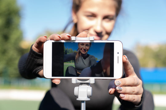Woman in sportswear vlogger sitting on artificial turf outdoor, demonstrating exercises for her online blog, records on smartphone camera on tripod. Broadcasting lesson via social media. 