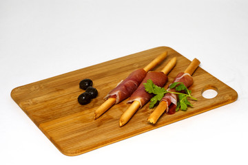 Bamboo board with bread sticks wrapped in ham. Olives and parsley.