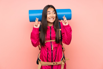 Young mountaineer Indian girl with a big backpack isolated on pink background with fingers crossing