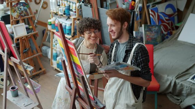 Man and woman cheerful young artists are talking looking at picture on easel discussing artwork in studio smiling enjoying arts class together