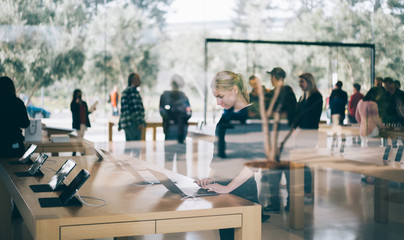 Young female customer standing near table with electronic products while examining the goods, store employees and visitors stand in blur on background. Millennial hipster girl checking device for buy - obrazy, fototapety, plakaty