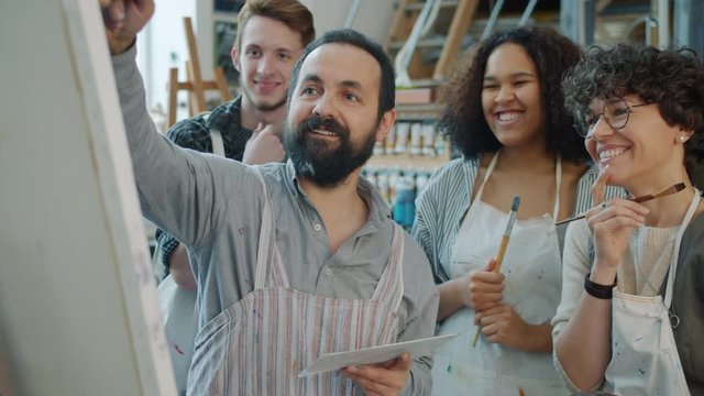 Slow motion of skilled middle-aged artist painting picture and talking to multi-ethnic group of students young people who are smiling and laughing