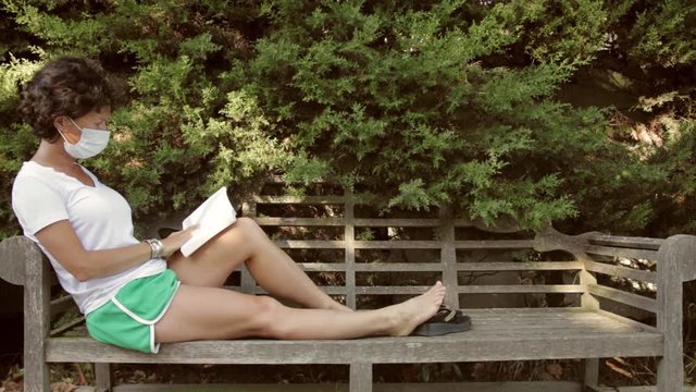 Une jeune femme assise sur un banc dans un parc lit un livre. Elle porte un masque.
