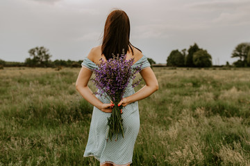 The girl holds in her hands a large bouquet of lilac and violet flowers behind back. In the background is green yellow grass and trees. Blue turquoise polka dot dress on a tanned brunette girl.