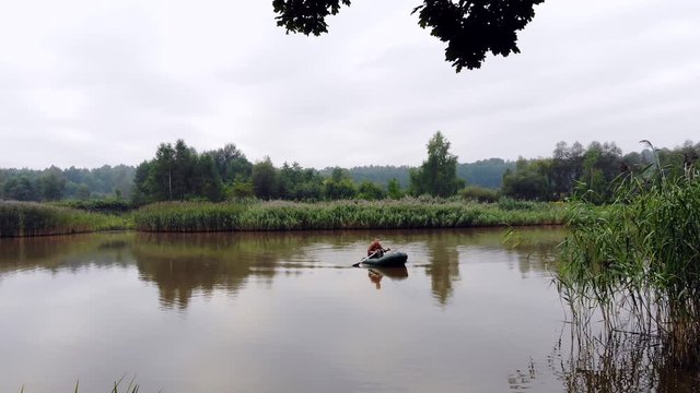 young man on an inflatable boat sits and throws a spinning or fishing rod into the water in the middle a lake or river. background of reeds and forest. Hipster guy fisherman active recreation, camping