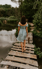 Fototapeta na wymiar A tanned brunette girl in a turquoise dress with white polka dots walks across the river alone an old wooden bridge. Against background of dark green trees. cloudy weather. back view. Woman at nature