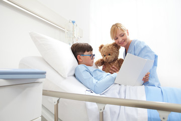 Child showing a book to nurse holding a teddy bear sitting beside him while lying in bed in hospital room