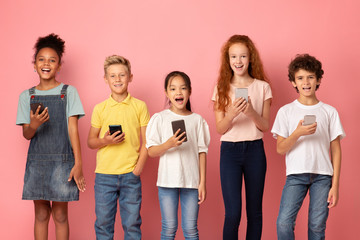 Fototapeta premium Online education. Diverse schoolchildren with mobile phones posing on pink background