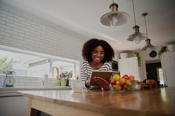 Cheerful young african woman browsing internet on digital tablet for new recipes in modern kitchen