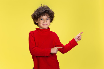 Pointing. Portrait of pretty young curly boy in red wear on yellow studio background. Childhood, expression, education, fun concept. Preschooler with bright facial expression and sincere emotions.