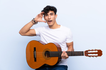 Young Argentinian man with guitar over isolated blue background with surprise and shocked facial expression