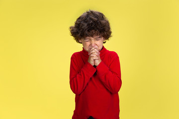 Fototapeta na wymiar Praying. Portrait of pretty young curly boy in red wear on yellow studio background. Childhood, expression, education, fun concept. Preschooler with bright facial expression and sincere emotions.