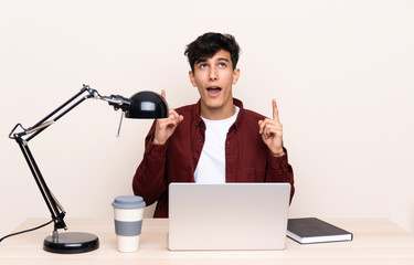 Young Argentinian man in a table with a laptop in his workplace pointing with the index finger a great idea