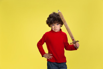 Warrior. Portrait of pretty young curly boy in red wear on yellow studio background. Childhood, expression, education, fun concept. Preschooler with bright facial expression and sincere emotions.