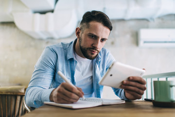 Focused male journalist using tablet for writing article in notebook while sitting in cafe