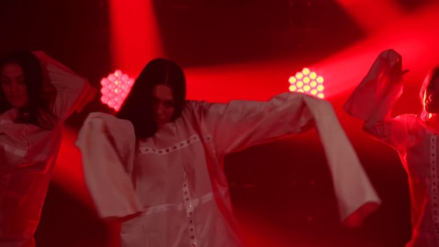 Three charming womans in white shirt are dancing on the stage. Shot in a dark studio with smoke and neon lighting. Red neon light effects. Dance show.