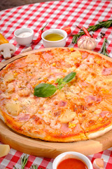 Hawaiian Italian pizza with parmesan cheese, pineapple, corn, ham toppings, fresh basil and tomatoes sauce on a wooden board, rustic village background, top view. European cuisine
