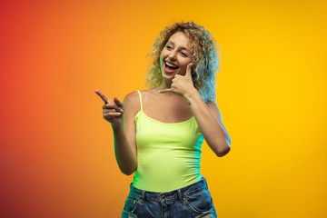 Need you to call. Caucasian young woman's portrait isolated on gradient studio background in neon. Beautiful female curly model in casual style. Concept of human emotions, facial expression, youth