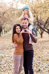 Mom, dad and their little daughter are posing in the autumn park. The girl sits on the shoulders of dad
