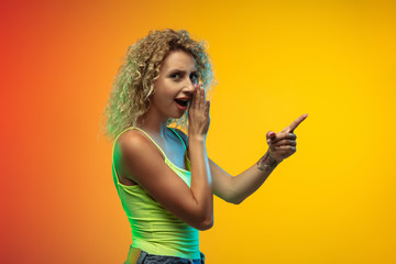 Pointing, showing. Caucasian young woman's portrait isolated on gradient studio background in neon. Beautiful female curly model in casual style. Concept of human emotions, facial expression, youth