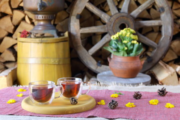 A couple glass mugs of black tea on the background of wooden wheel, yellow tub and stack of firewood.