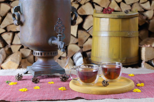 A Couple Glass Mugs Of Black Tea On The Background Of Samovar, Yellow Tub And Stack Of Firewood.