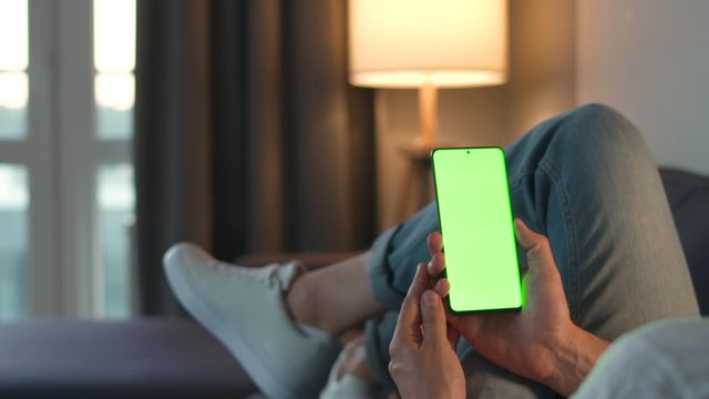 Man at home lying on a sofa and using smartphone with green mock-up screen in vertical mode. He browsing Internet, watching content, videos, blogs. POV.